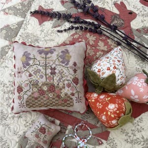 May include: A decorative pillow with a cross-stitch floral design, featuring a basket of flowers and birds, with a red border. Accompanying the pillow are fabric strawberries in various floral patterns and a sprig of lavender.
