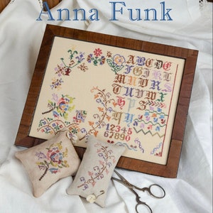 May include: A framed cross-stitch sampler with a floral design and the alphabet and numbers embroidered in colorful thread. The sampler is displayed on a white fabric surface with two small cross-stitch pillows and a pair of antique scissors. The text "Anna Funk" is displayed above the frame and "Erica Michaels Needleart Designs" is displayed below the frame.