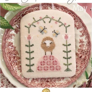 May include: Cross-stitch embroidery featuring a sheep in a pink dress, framed by green floral elements and butterflies. The text "The Shepherd's Year" and "May" are visible. The design is on a cream-colored fabric, with the "October House Fiber Arts" logo.