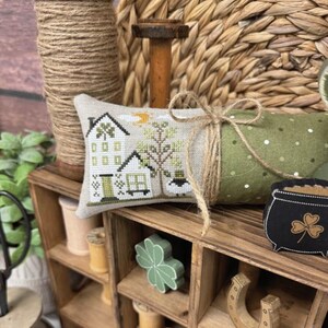 May include: A decorative display featuring a cross-stitch pillow with a house design, a green polka-dot fabric roll, and a black pot with a shamrock. The display includes the text "EWE HAPPY HOME" and "PRIMROSE COTTAGE". Other items include a horseshoe and a wooden spool.