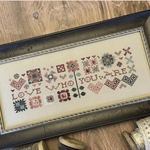 May include: A framed cross-stitch sampler with a white background and colorful embroidery. The sampler features a variety of small quilt blocks and the text "Love who you are."