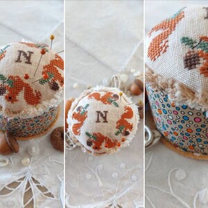 May include: A white fabric pincushion with a brown and orange embroidered design of squirrels and acorns. The pincushion is round and has a brown and white polka dot fabric base. The pincushion is decorated with the letter "N".