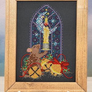 May include: A cross-stitch embroidery in a light wood frame featuring a brown mouse holding a lit candle in front of a stained-glass window design with a crescent moon, surrounded by yellow bells, red ribbons, and green holly on a black fabric.