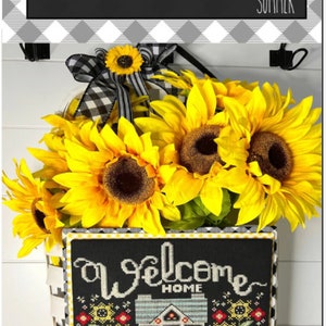 May include: A cross-stitch pattern featuring a house with sunflowers and the text "Welcome Home" on a black background. The pattern is framed with a black and white checkered border and the text "Stitching with the Housewives" is at the top.