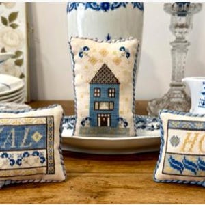 May include: Three rectangular and vertical fabric pillows featuring intricate cross-stitch embroidery in shades of blue, cream, and gold. Two pillows display the words HEAL and HOME, while the central vertical pillow depicts a blue house.