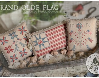 May include: Three decorative, stitched pillows with patriotic designs. One pillow features a red and white striped flag, another has blue and red starbursts, and the third displays a tree with flags. The pillows are in a metal tray with buttons and fabric scraps. The text "GRAND OLDE FLAG" is visible.