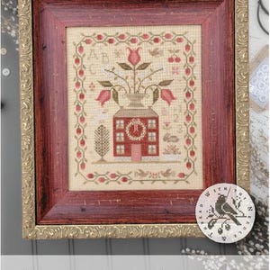 May include: Framed cross-stitch sampler with a red house, flowers, and the text "ABC ~ 123 SAMPLER." The sampler is in a red frame with a gold-colored ornate outer frame. The design includes birds and floral elements.