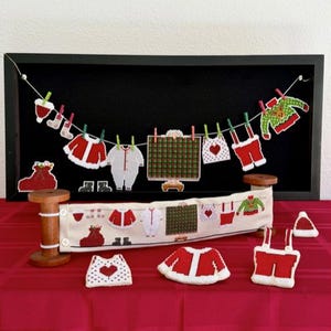 May include: A Christmas-themed display featuring miniature, handcrafted clothing items. A clothesline with tiny outfits hangs against a black backdrop. Additional pieces, including a sweater, onesie, and pants, are arranged on a red tablecloth, creating a festive scene.
