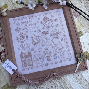 May include: A framed cross-stitch sampler with a brown wooden frame. The sampler features a variety of embroidered motifs, including a peacock, flowers, a house, and the alphabet. The color palette is primarily shades of brown and white. A pair of scissors and a tag are also visible.