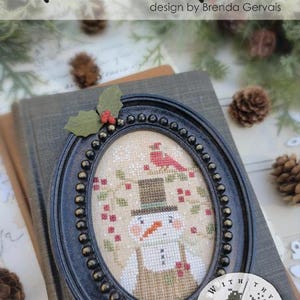 May include: An oval black beaded frame containing a cross-stitch pattern of a snowman wearing a top hat, with a red cardinal and holly berries. The text reads #CS386 Pixie Pics FREEZIN' FROSTY design by Brenda Gervais.