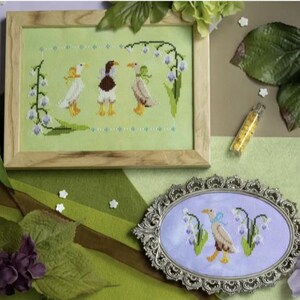 May include: Cross-stitch art featuring three ducks with floral accents in a wooden frame. The ducks are in various colors, with yellow feet and beaks. A second, smaller cross-stitch piece in an oval frame is also visible, along with decorative elements.