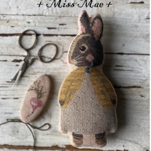 May include: A brown and white bunny doll with a yellow jacket and a blue ribbon around its neck. The doll is sitting on a white wooden surface. The text "Animal Crackers Series + Miss Mae +" is at the top of the image and the text "Stacy Nash Designs" is at the bottom of the image.