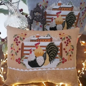 May include: A beige pillow with a white scalloped edge features a cross-stitch design of two roosters in a snowy winter scene. The roosters are standing in front of a red barn with a white roof. The scene is surrounded by holly branches with red berries.