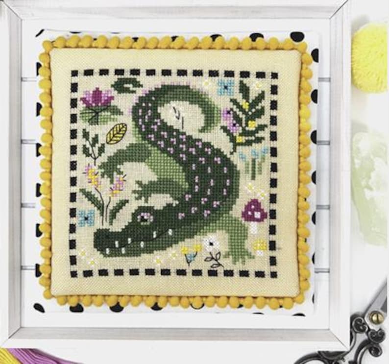Pu&ograve; includere: Un ricamo a punto croce che rappresenta un alligatore verde con macchie viola, circondato da fiori e funghi colorati. Il disegno &egrave; incorniciato da un bordo a scacchi bianco e nero e da una bordura di pon pon gialli.