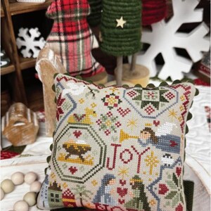 May include: A decorative pillow with a cross-stitch design featuring the word "JOY" and Nativity scene elements. The pillow has a beige background with red, green, and blue accents. The text "NATIVITY QUAKER" and "PRIMROSE COTTAGE" are visible.