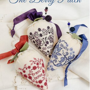 May include: Three embroidered fabric strawberry pincushions with different designs and colors. The pincushions are on a white fabric background. The text "The Berry Patch" is at the top of the image. The text "ERICA MICHAELS NEEDLEART DESIGNS" is at the bottom of the image.