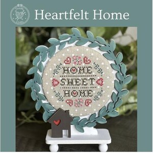 May include: A white wooden stand with a green leafy wreath frame holding a cross-stitch embroidery with the text "Home Sweet Home" in red and black lettering. The embroidery is surrounded by white polka dots and red hearts. A small brown wooden house with a red heart on the door is in front of the stand.