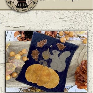 May include: Wool applique craft kit featuring an autumn squirrel design. The design includes a pumpkin, leaves, and an acorn on a navy blue background. The finished size is 9 x 11 inches.