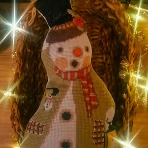 May include: A cross-stitched snowman ornament wearing a black top hat and a red and white striped scarf. The snowman has a tan coat, a small orange nose, and black button details. A tiny snowman figure and a yellow lantern are stitched on the coat.
