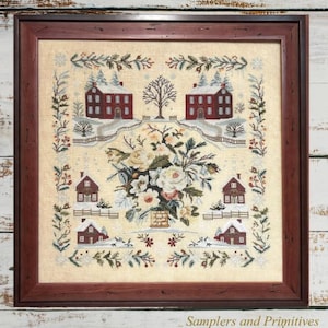May include: A framed cross-stitch sampler on beige fabric featuring a central floral arrangement surrounded by six red houses in a snowy landscape. The border consists of green pine branches and red berries. The text Samplers and Primitives is visible.
