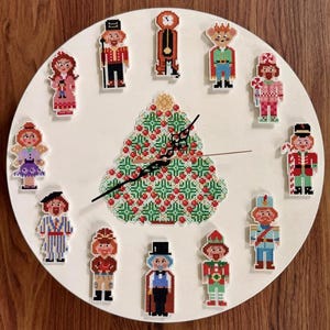 May include: A round white wall clock featuring a central cross-stitch pattern of a green and red Christmas tree. Twelve individual cross-stitch figures representing various characters are arranged around the dial as hour markers, with black hands.