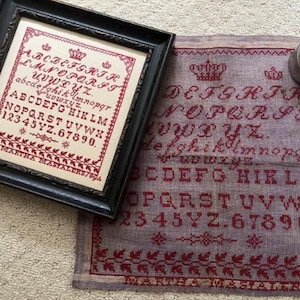 May include: Framed antique sampler with a black wooden frame. The cross-stitch design features the alphabet, numbers, and decorative elements in red thread on a cream-colored fabric. A second, unframed sampler is also visible.