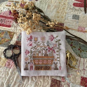 May include: A cross-stitch embroidery featuring a spring garden scene with flowers, bunnies, and the word "SPRING". The embroidery is on a light-colored fabric, with a pair of vintage scissors and dried flowers nearby.