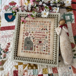 May include: A framed cross-stitch sampler with a house, flowers, and the alphabet in a cream-colored frame with a beaded border. A white angel figurine with floral embroidery is on the right side of the image.