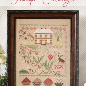 May include: A framed cross-stitch embroidery featuring a cottage with a snowy roof, surrounded by tulips and a rabbit. The alphabet is stitched around the cottage and tulips, and there are three baskets of strawberries at the bottom. The title "Tulip Cottage" is stitched above the frame.