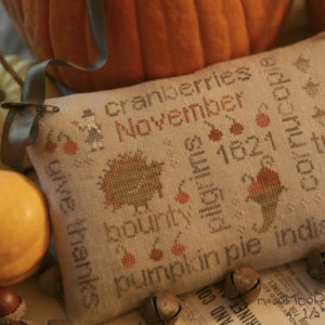 May include: A beige cross-stitch pillow with a Thanksgiving theme. The pillow features the words "cranberries", "November", "give thanks", "bounty", "1621", "corn", "turkey", "pumpkin pie", and "indian corn".