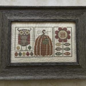 May include: Framed cross-stitch artwork featuring an owl, pumpkin, and flower. The owl is brown and red, the pumpkin is brown and green, and the flower is red and brown. The frame is gray.