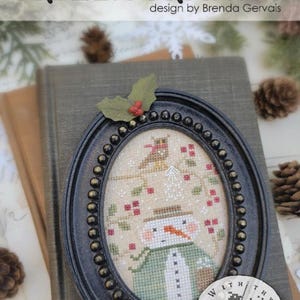 May include: An oval black frame with beaded detailing contains a cross-stitch pattern of a snowman wearing a green coat and hat, with an owl perched on a branch above. The text reads #CS385 Pixie Pics FROZEN FRANNY design by Brenda Gervais.