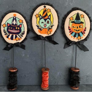 May include: Three round cross-stitch ornaments on thin black rods with wooden spool bases. The left features a black cat, the center a vampire cat, and the right a jack-o'-lantern, all with black fur trim and black ribbon bows at the base.