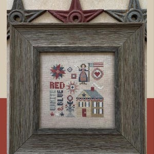 May include: A framed cross-stitch sampler with a patriotic theme. The design features a house, stars, and the words "RED, WHITE & BLUE." The frame is a weathered gray, and the sampler is displayed against a neutral background.