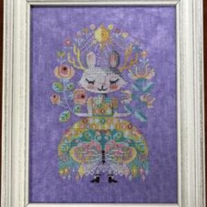 May include: A framed cross-stitch artwork featuring a whimsical deer-like character with closed eyes, adorned with floral and geometric patterns. The deer wears a colorful dress and is framed in a white wooden frame. The background is a solid lavender color.