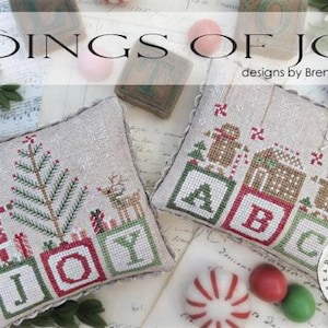 May include: Two square linen pillows featuring cross-stitch embroidery. One pillow displays a Christmas tree, a reindeer, and the letters J, O, Y. The other shows gingerbread figures, a house, and the letters A, B, C. Text reads TIDINGS OF JOY #CS389.