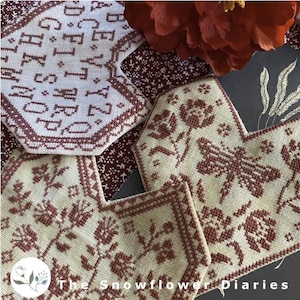 May include: Close-up of several cross-stitch samplers with floral and alphabet designs in shades of cream and burgundy. The samplers are in various shapes, including a hexagon and a heart. The text "The Snowflower Diaries" is visible in the bottom left corner.