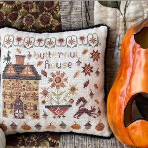 May include: A white pillow with a cross-stitch design of a house with the text "butternut house". The pillow is on a brown and green quilt with a pumpkin and a jack-o'-lantern.