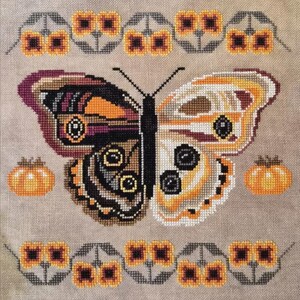 May include: A cross-stitch embroidery on beige fabric featuring a large butterfly with a dark left wing and a light right wing. The design includes two orange pumpkins on the sides and horizontal borders of small yellow flowers with gray stems.