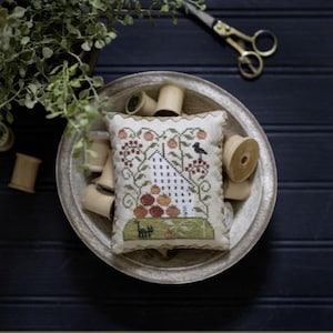 May include: A white and brown cross-stitch sampler with a house, trees, and birds. The sampler is in a silver bowl with wooden spools of thread. The title of the sampler is "Tomato Hill" and the text "Plum Street Samplers" is in the top right corner.