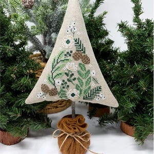 May include: A decorative, handmade Christmas tree with a beige fabric cone, embroidered with white flowers, green leaves, and brown pinecones. The tree is mounted on a wooden stick and a brown felt base, tied with twine. The background features miniature Christmas trees.