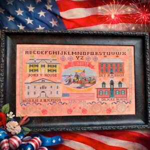 May include: A framed cross-stitch sampler with an American patriotic theme. The sampler features illustrations of historic buildings, the word "LIBERTY", and the American flag. The background is a peach color with floral accents. The frame is black.