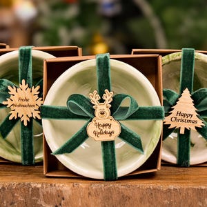 Op de afbeelding: Een verzameling kleine, decoratieve kommen in kartonnen dozen, vastgebonden met groen fluwelen lint. Elke kom heeft een houten label met feestelijke groeten zoals "Merry Christmas" en "Happy Holidays". De kommen zijn lichtgroen met een marmerpatroon.