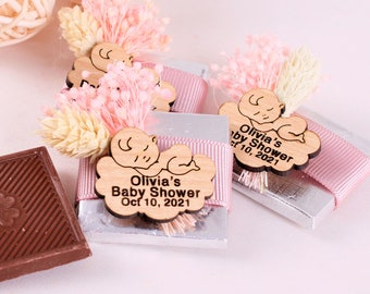 May include: Two small chocolate bars wrapped in silver foil with pink ribbon and a wooden tag with the text "Olivia's Baby Shower Oct 10, 2021" and a small wooden baby figure. The tag is attached to a small bouquet of dried flowers.