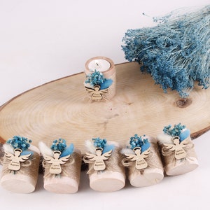 Pu&ograve; includere: Sette piccoli cilindri di legno avvolti in juta e decorati con fiori secchi blu e bianchi. Ogni cilindro ha un ritaglio di angelo in legno con il testo "Battesimo di Liam" su di esso. Un cilindro pi&ugrave; grande con una candela &egrave; sullo sfondo.