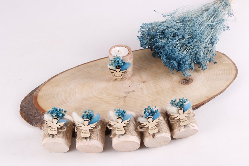 Pu&ograve; includere: Sette piccoli cilindri di legno avvolti in juta e decorati con fiori secchi blu e bianchi. Ogni cilindro ha un ritaglio di angelo in legno con il testo "Battesimo di Liam" su di esso. Un cilindro pi&ugrave; grande con una candela &egrave; sullo sfondo.
