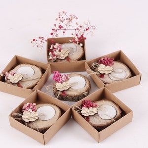 May include: Six small, brown cardboard boxes each contain a wooden candle holder with a small white candle. Each holder is decorated with a small bouquet of pink dried flowers and a personalized wooden tag.