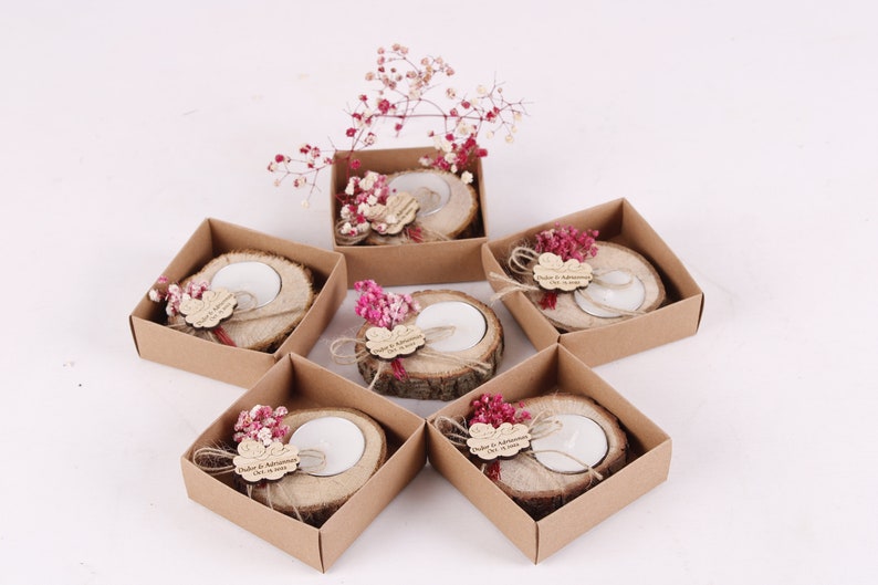 May include: Six small, brown cardboard boxes each contain a wooden candle holder with a small white candle. Each holder is decorated with a small bouquet of pink dried flowers and a personalized wooden tag.