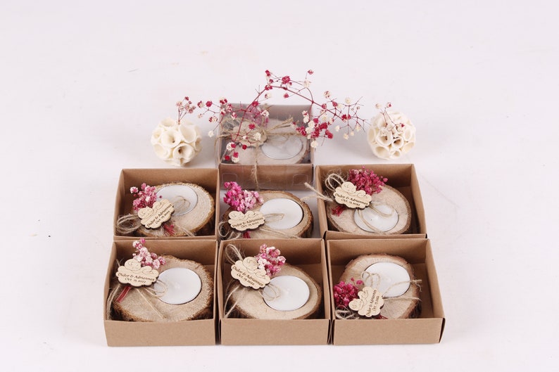 May include: Six small brown boxes each contain a wooden candle holder with a small white candle, a small sprig of dried pink flowers, and a personalized wooden tag. The boxes are arranged in two rows of three.