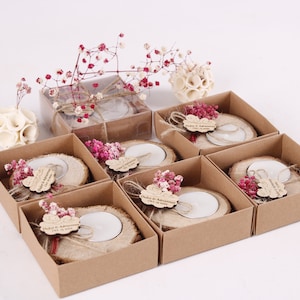 May include: Set of eight small, brown cardboard boxes with lids, each containing a wooden candle holder with a small white candle. The candle holders are decorated with dried pink flowers and a small wooden tag with the text "Duket & Adhmiransa 1.12.2023".